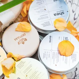 Zestaw kosmetyków naturalnych w szklanym naczyniu, kremy shea w metalowych puszkach z drewnianymi wieczkami, ozdobione płatkami kwiatów i źdźbłem trawy.