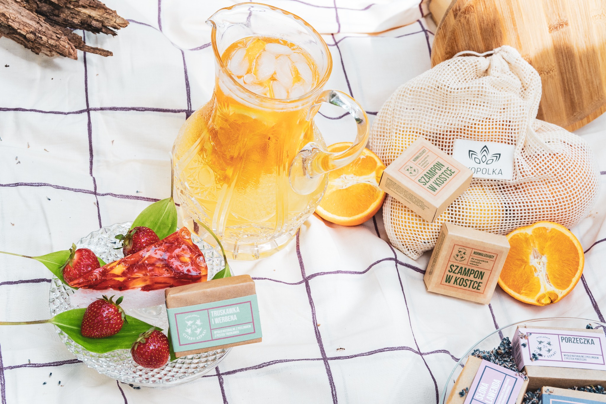 Kryształowy dzbanek z lemoniadą i lodem na piknikowym kocu, otoczony kostkami mydła i szamponu w papierowych opakowaniach, pomarańczami i deserem truskawkowym na szklanym talerzu.