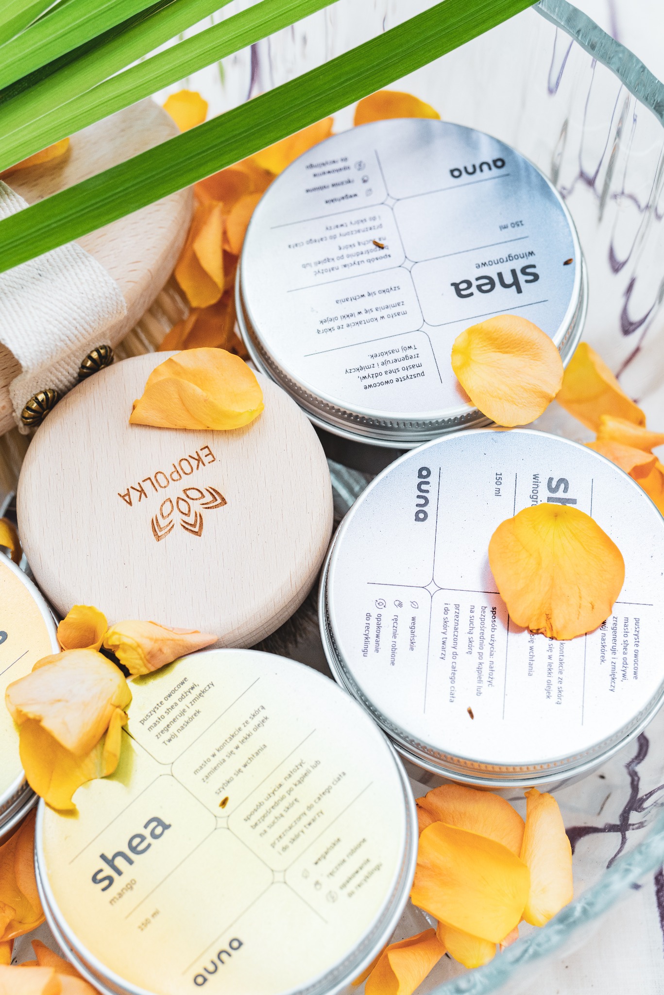 Zestaw kosmetyków naturalnych w szklanym naczyniu, kremy shea w metalowych puszkach z drewnianymi wieczkami, ozdobione płatkami kwiatów i źdźbłem trawy.