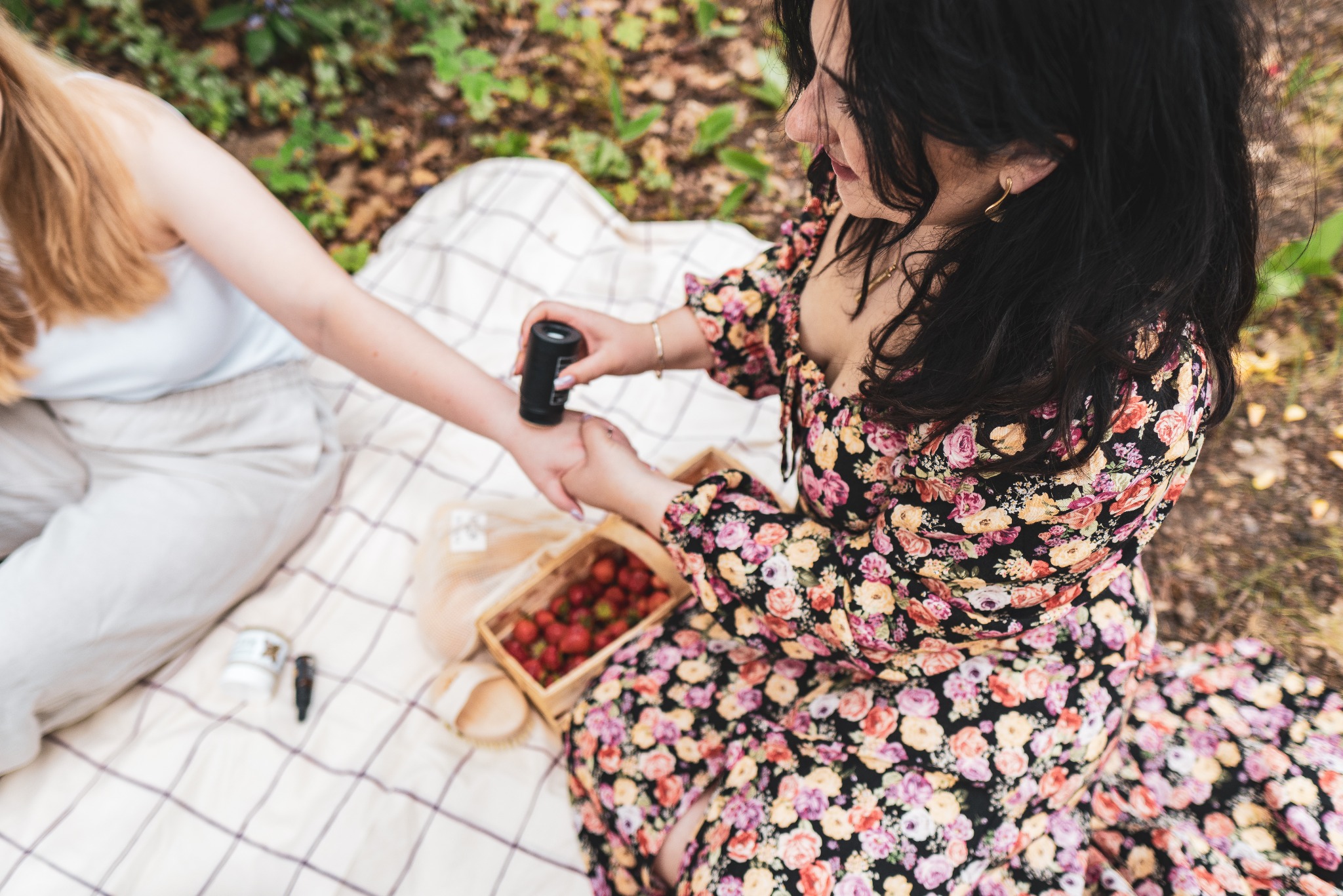 Kobieta w kwiecistej sukience aplikuje kosmetyk na przedramię drugiej kobiety, siedząc na kocu piknikowym obok drewnianego koszyka z truskawkami.
