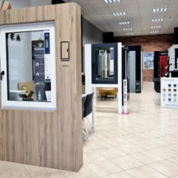 Ekspozycja okien w salonie sprzedaży, prezentacja różnych modeli i kolorów ram okiennych. Widoczny panel z próbkami kolorów Eurocolor oraz okno z naklejką 'Livio'.