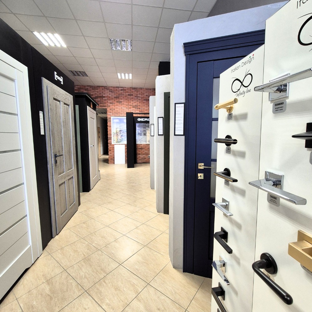 Ekspozycja różnych modeli drzwi wewnętrznych w salonie, prezentacja klamek i okuć, widok na korytarz z płytkami podłogowymi i ceglaną ścianą w tle.