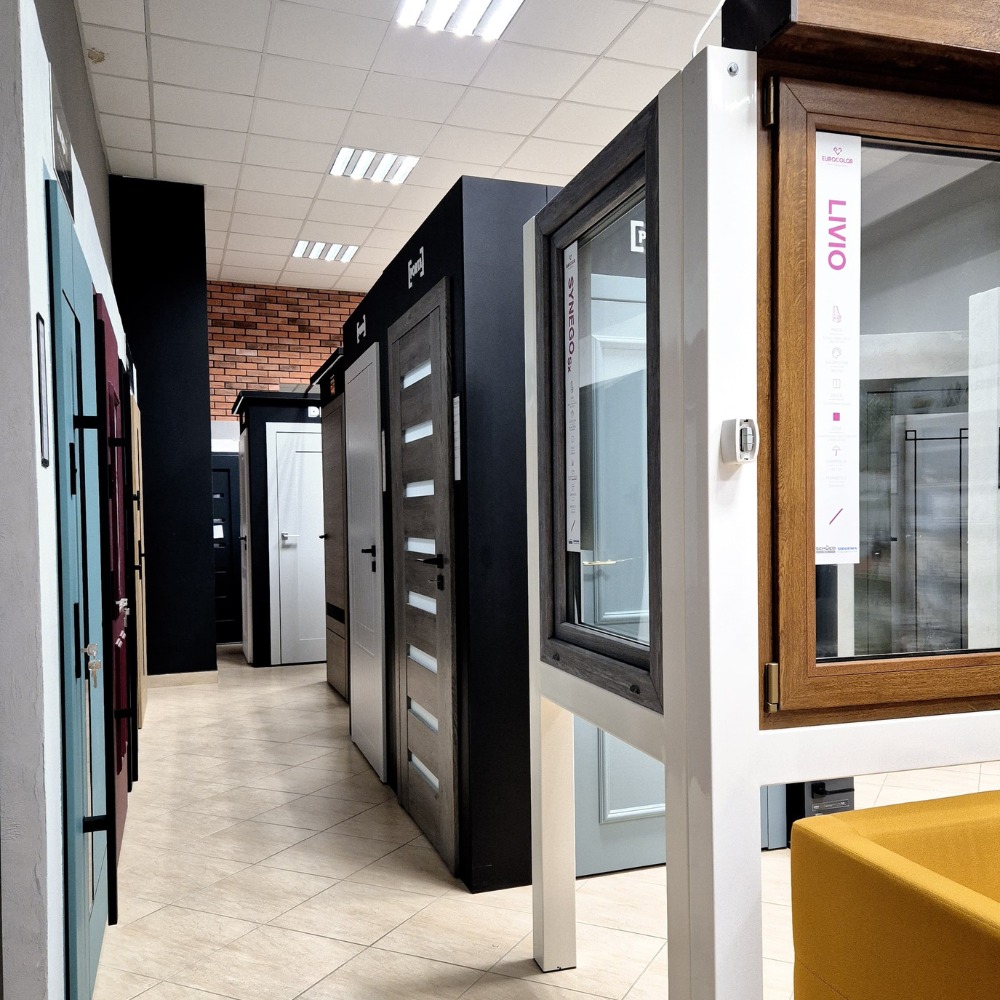 Ekspozycja różnych modeli drzwi i okien w salonie, prezentowanych w rzędzie z oświetleniem sufitowym i kafelkową podłogą.