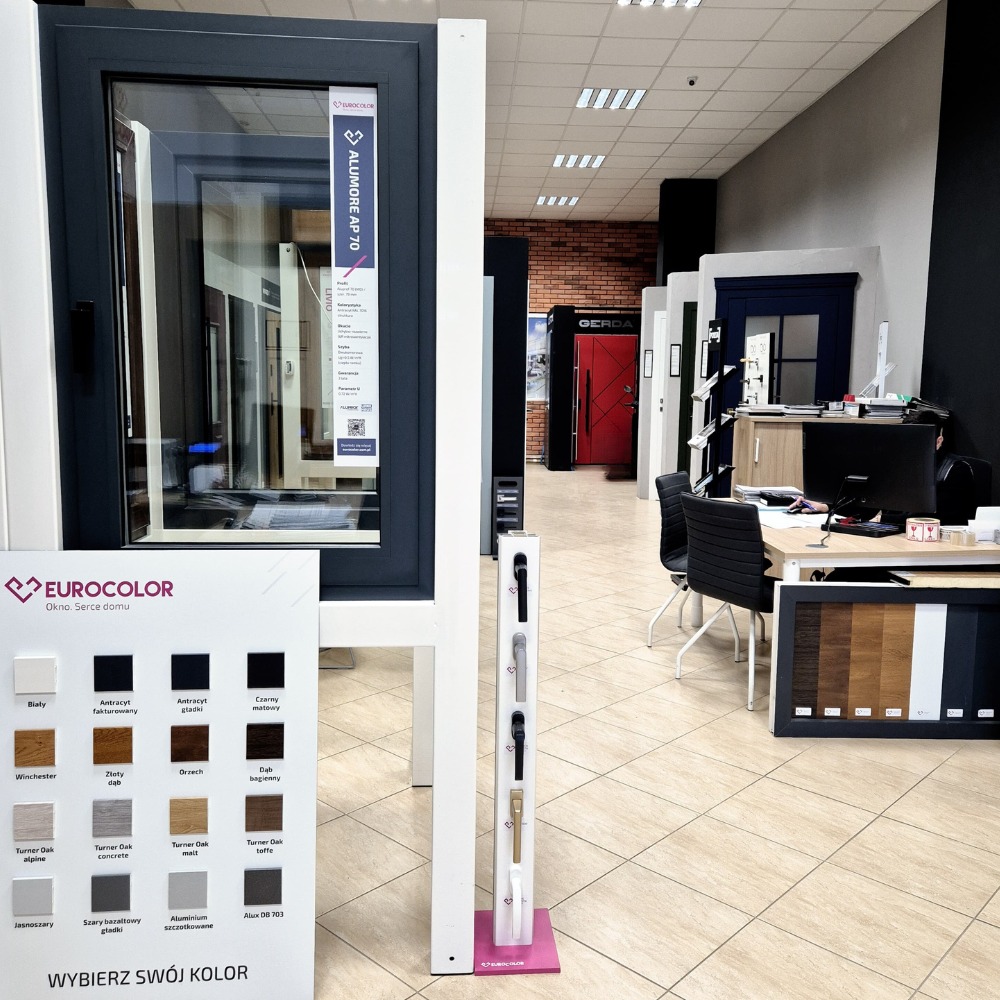 Ekspozycja okien PCV marki Eurocolor w salonie sprzedaży, prezentująca różnorodność kolorów i modeli, z paletą barw do wyboru i wzornikiem klamek na pierwszym planie, w tle widoczne drzwi Gerda.