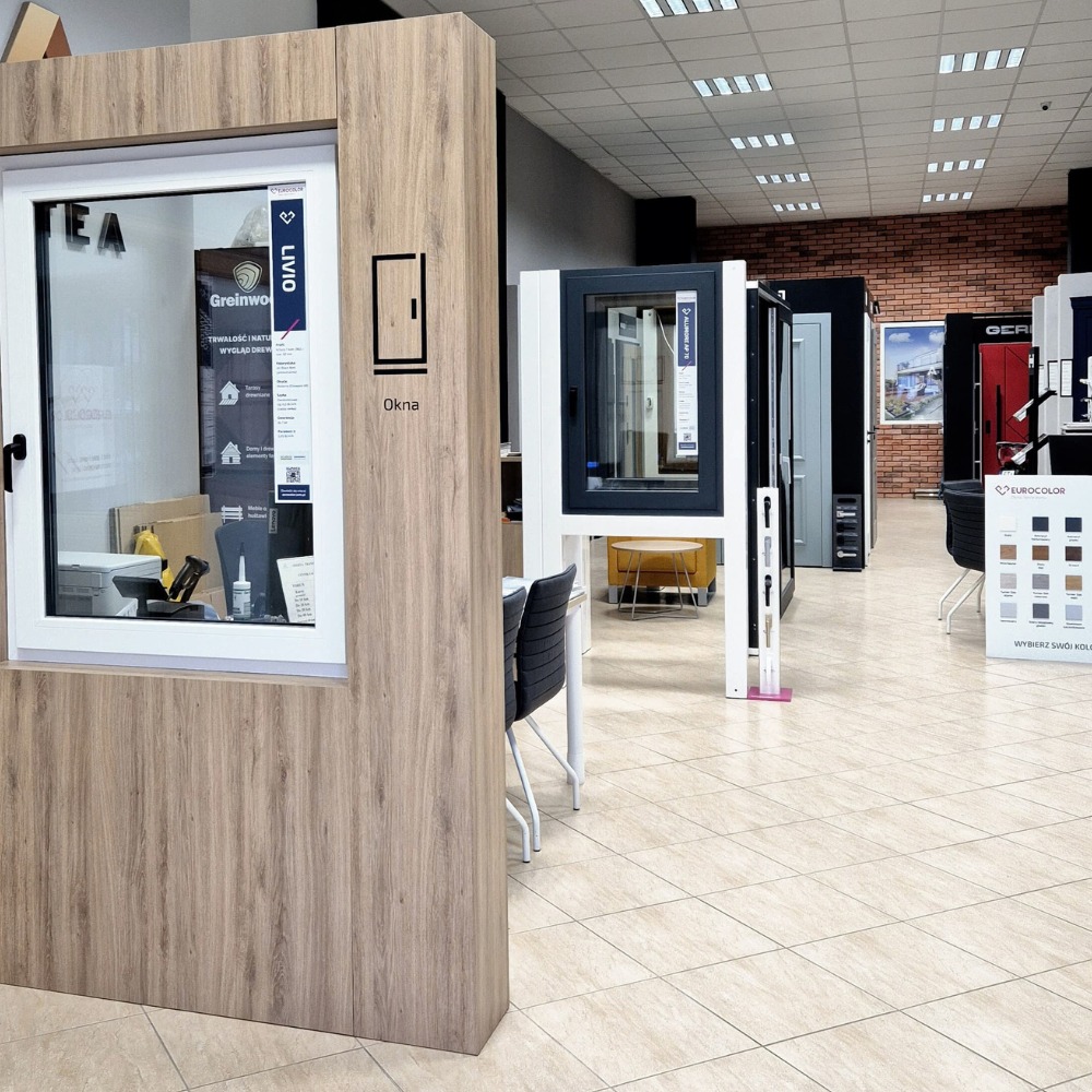 Ekspozycja okien w salonie sprzedaży, prezentacja różnych modeli i kolorów ram okiennych. Widoczny panel z próbkami kolorów Eurocolor oraz okno z naklejką 'Livio'.