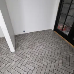 Ułożona szara podłoga w jodełkę z płytek ceramicznych w rogu pomieszczenia, widoczna czarna rama przeszklonych drzwi balkonowych i fragment białej ściany z puszką elektryczną.