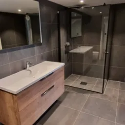 Nowoczesna łazienka z szarymi płytkami, drewnianą szafką z umywalką i kabiną prysznicową typu walk-in z czarnymi ramami.