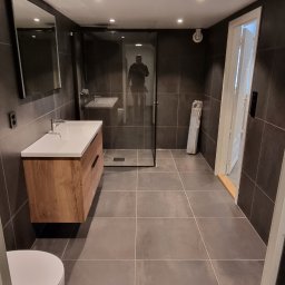 Dawid - Nowoczesna łazienka z szarymi płytkami, drewnianą szafką pod umywalką, kabiną prysznicową typu walk-in i lustrem na ścianie. W lustrze kabiny widoczne odbicie osoby fotografującej.