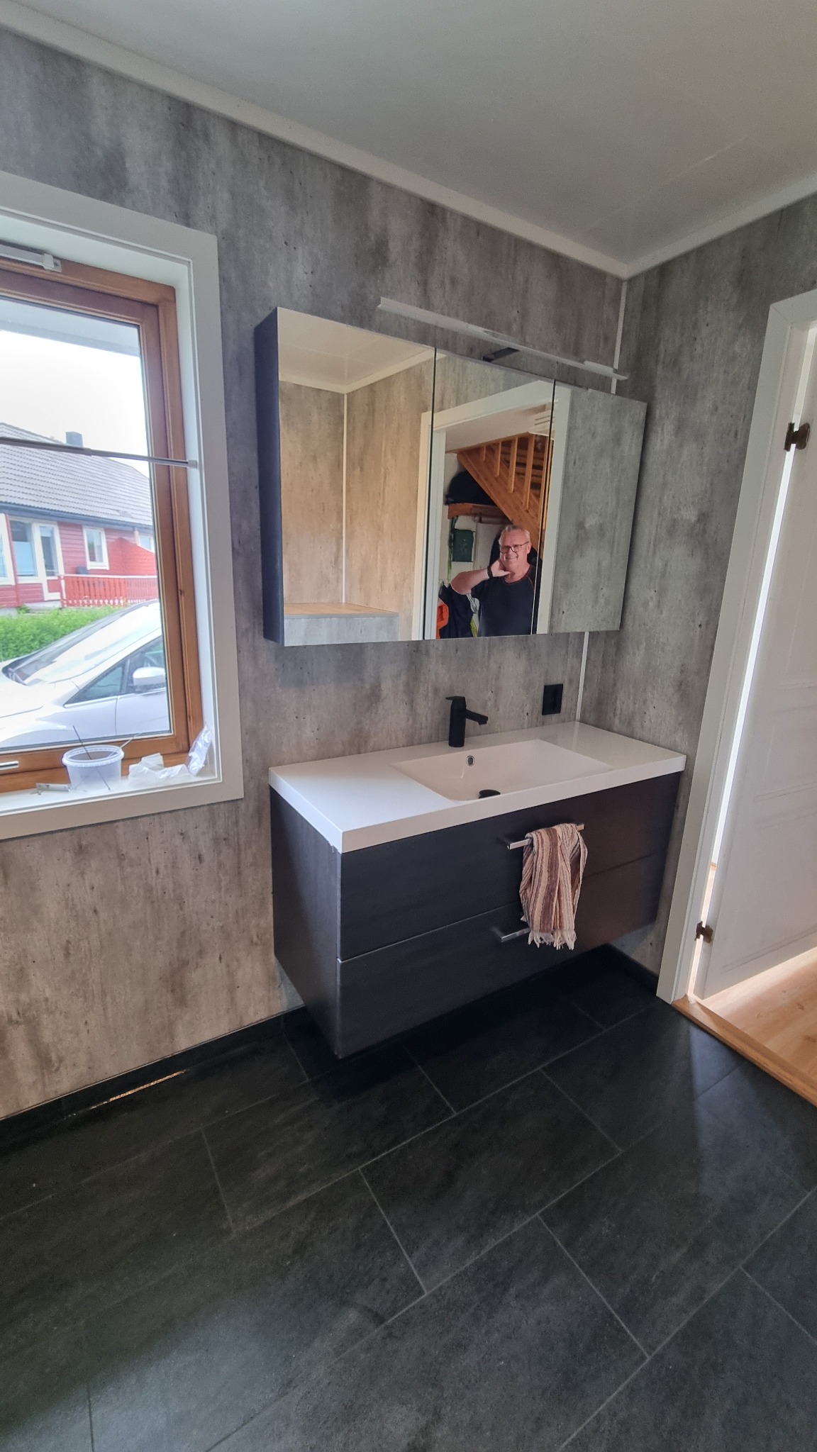 Nowoczesna łazienka w Augustowie z szarymi płytkami imitującymi beton na ścianach, ciemną szafką pod umywalką z białym blatem i lustrem z szafką, w którym odbija się osoba robiąca zdjęcie.