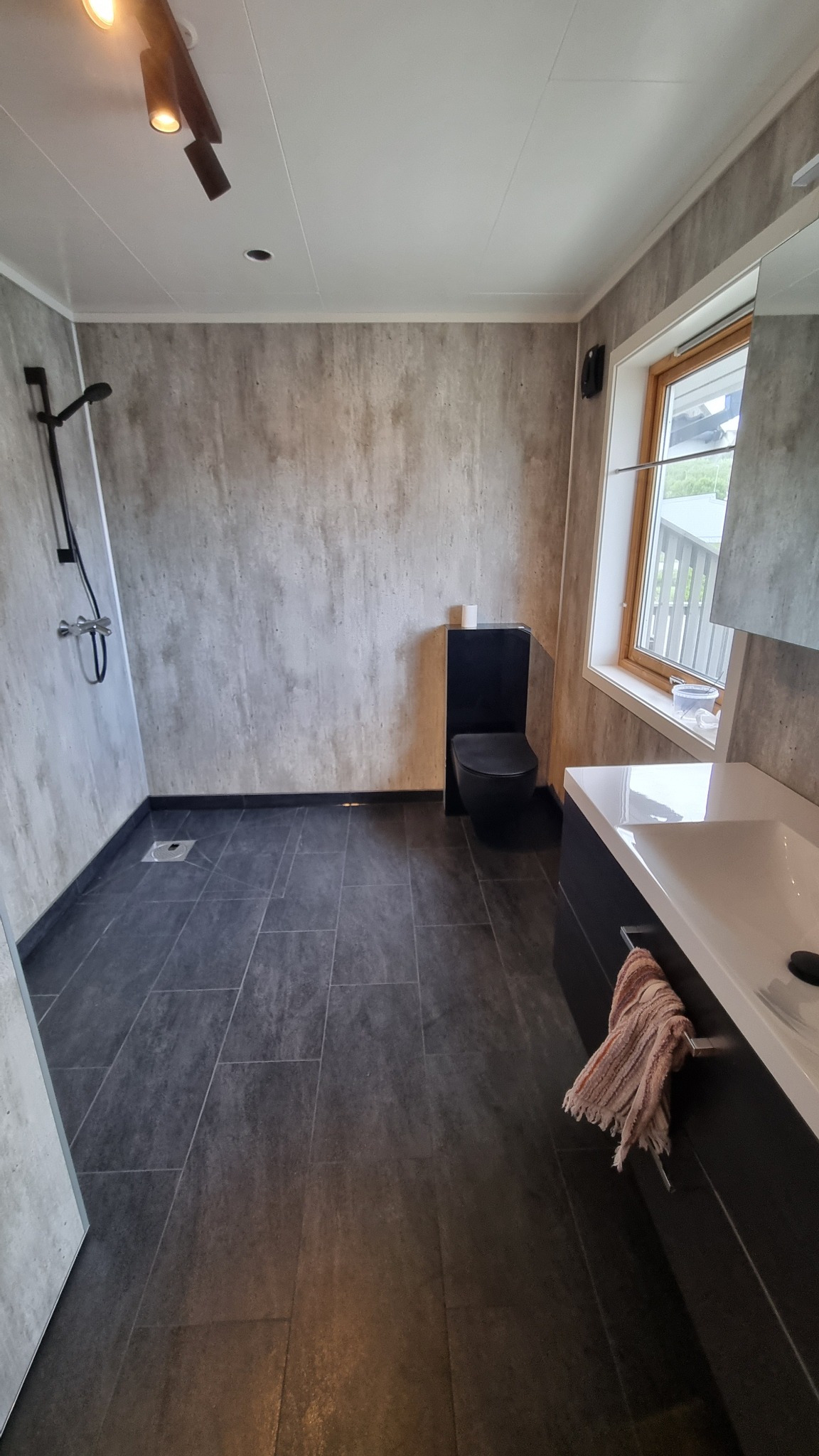 Nowoczesna łazienka z czarną toaletą typu Geberit, szarymi panelami ściennymi imitującymi beton, czarną podłogą z płytek i białym blatem umywalki z szufladami.