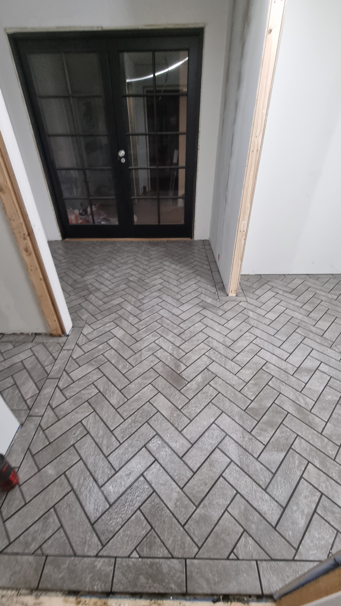 Ułożona szara podłoga z płytek w jodełkę, widoczne czarne, przeszklone drzwi dwuskrzydłowe i surowy stan ścian z widocznymi elementami konstrukcyjnymi.