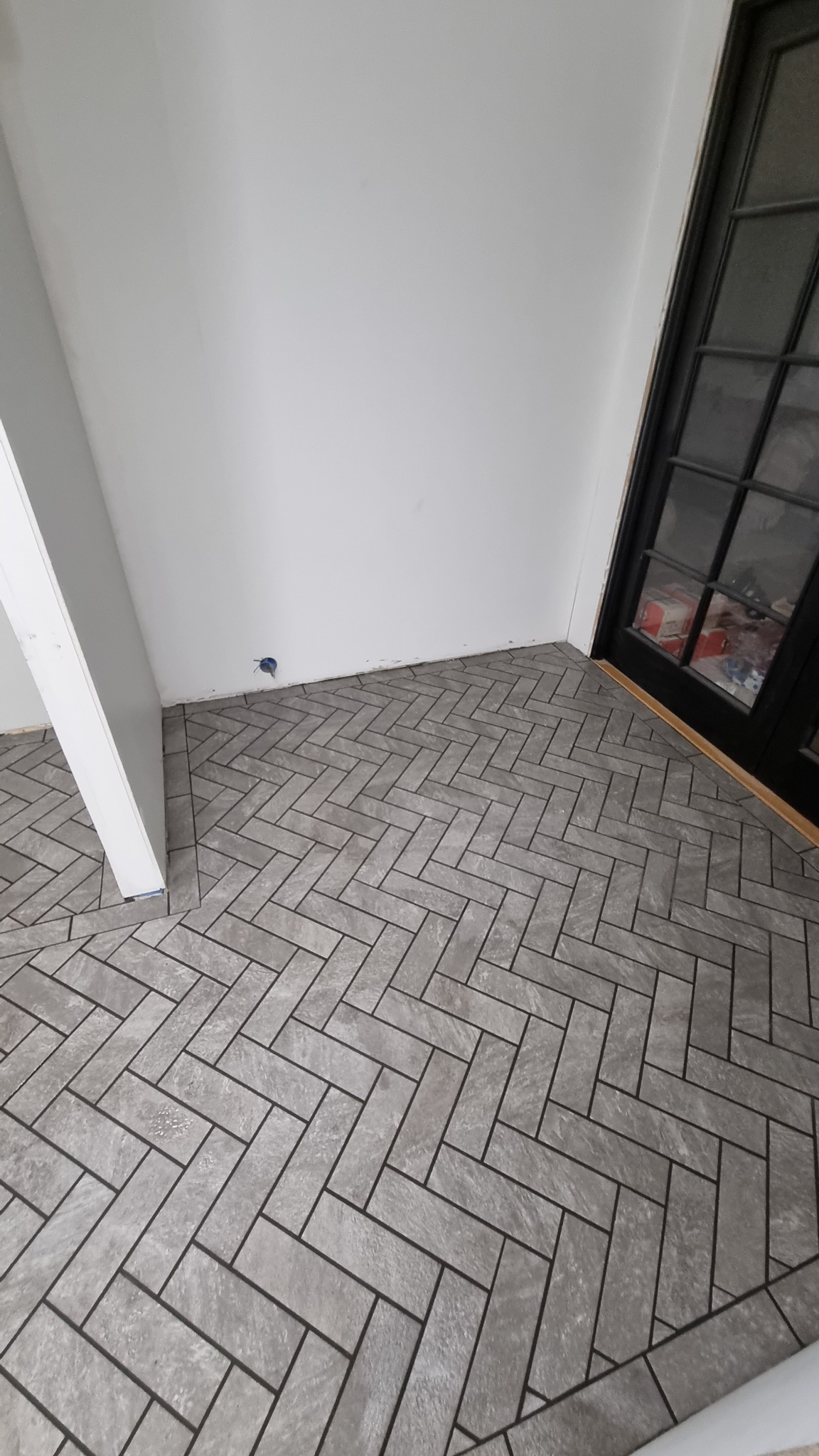 Ułożona szara podłoga w jodełkę z płytek ceramicznych w rogu pomieszczenia, widoczna czarna rama przeszklonych drzwi balkonowych i fragment białej ściany z puszką elektryczną.