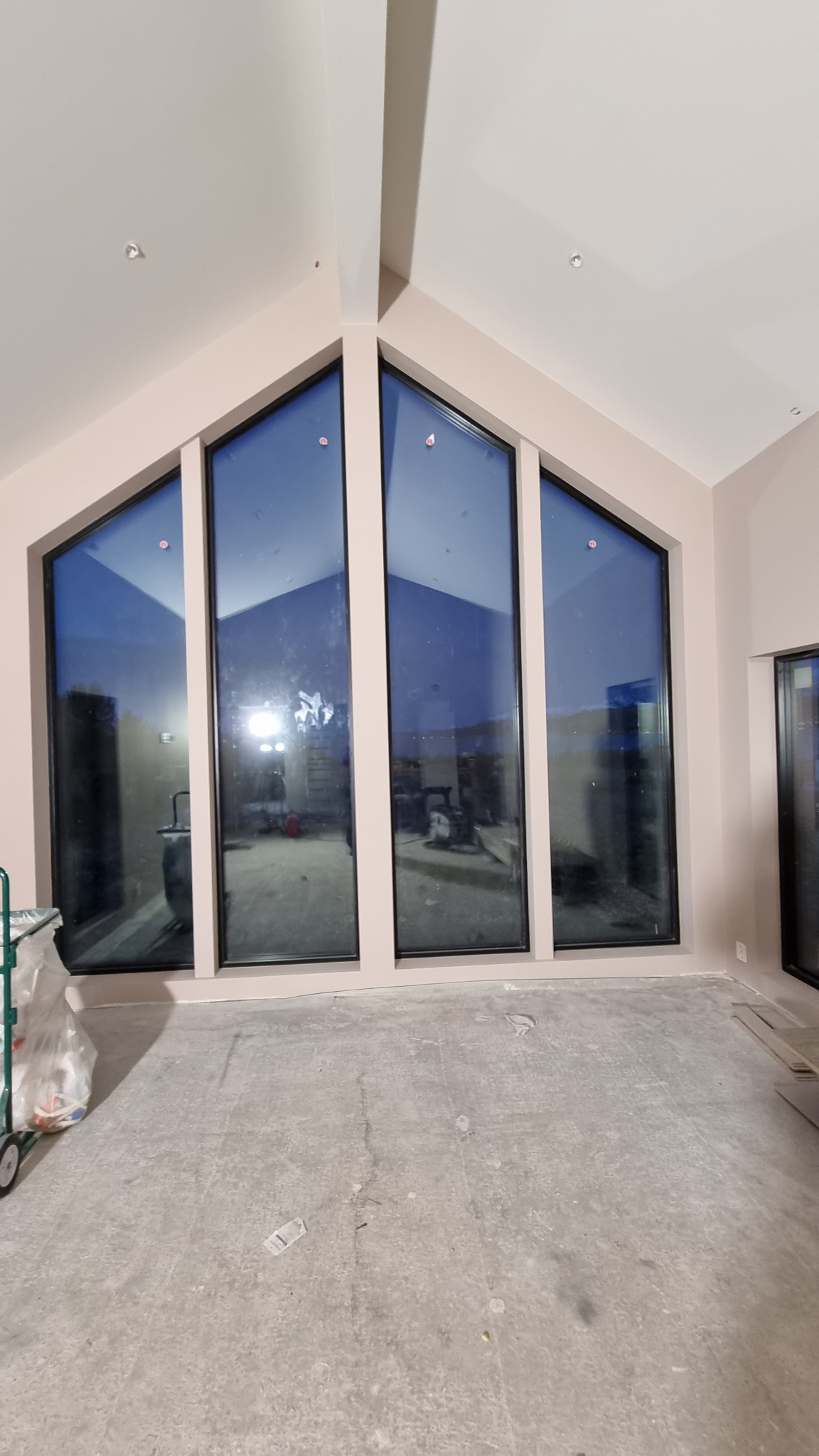Wnętrze z wysokimi trójkątnymi oknami w czarnych ramach, widok na zewnątrz częściowo zasłonięty przez sprzęt budowlany, surowy beton na podłodze przed wykończeniem.