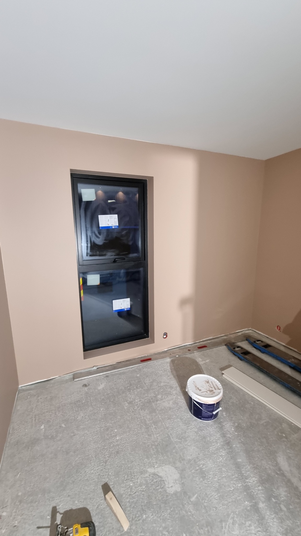 Pokój w trakcie remontu, ściany pomalowane na jasny beż, czarna rama okienna, na podłodze surowy beton, puszka z farbą, narzędzia do cięcia paneli i listwy przypodłogowe.