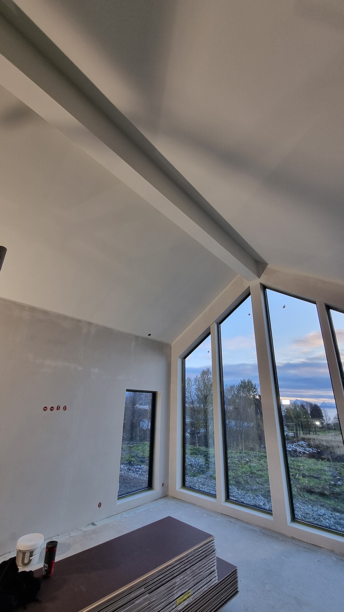 Wnętrze w trakcie prac wykończeniowych: surowa ściana z widocznymi puszkami elektrycznymi, okna z widokiem na krajobraz, stos płyt podłogowych i wiadro farby.
