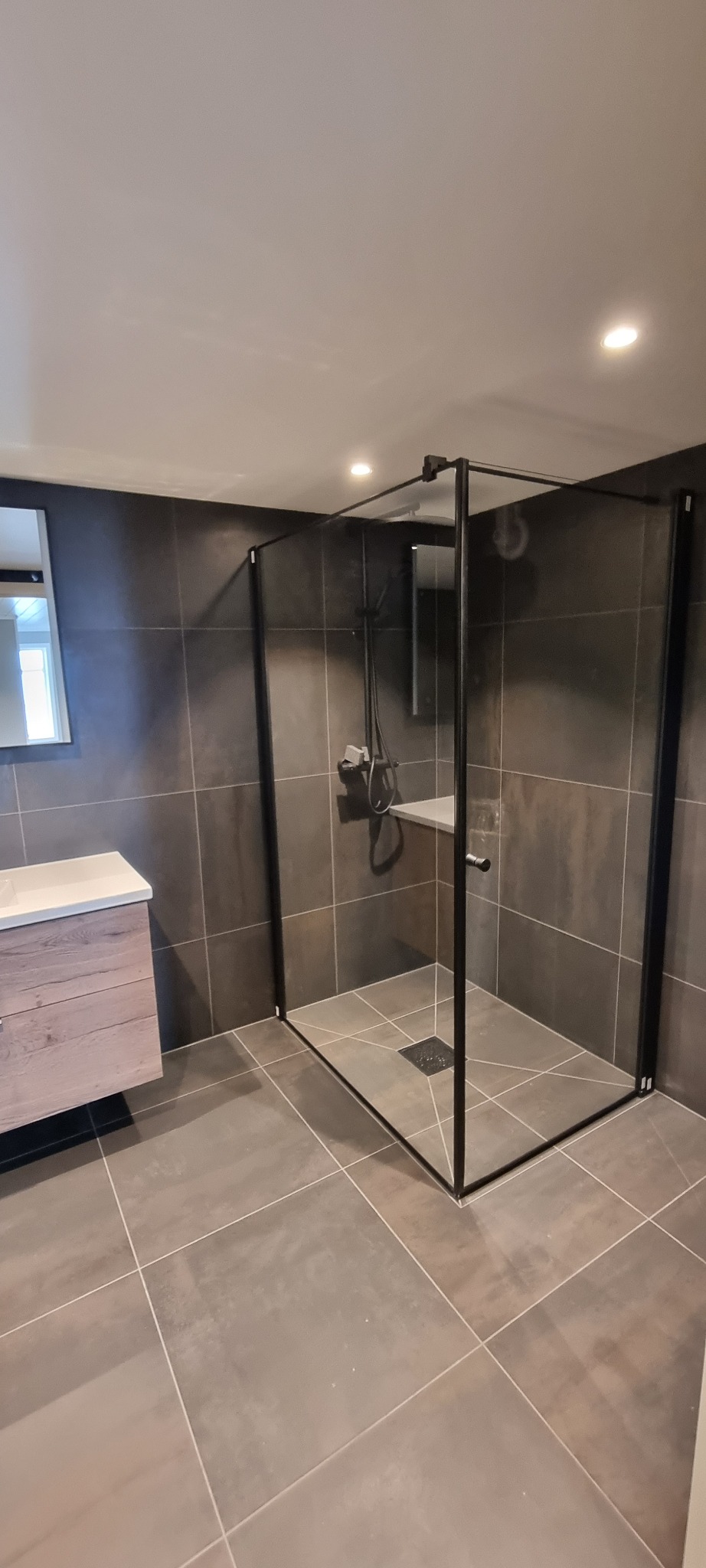 Nowoczesna łazienka z kabiną prysznicową typu walk-in z czarnymi profilami, szarymi płytkami na ścianach i podłodze oraz drewnianą szafką pod umywalką.