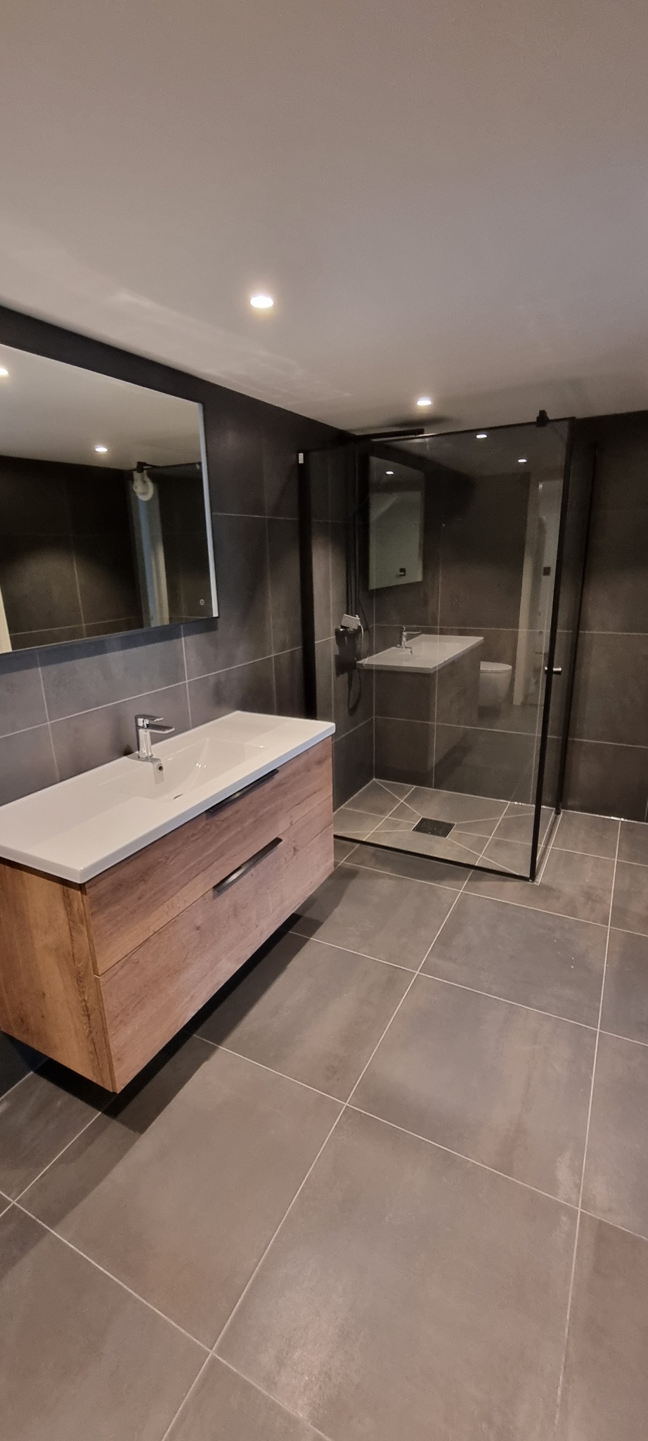 Nowoczesna łazienka z szarymi płytkami, drewnianą szafką z umywalką i kabiną prysznicową typu walk-in z czarnymi ramami.