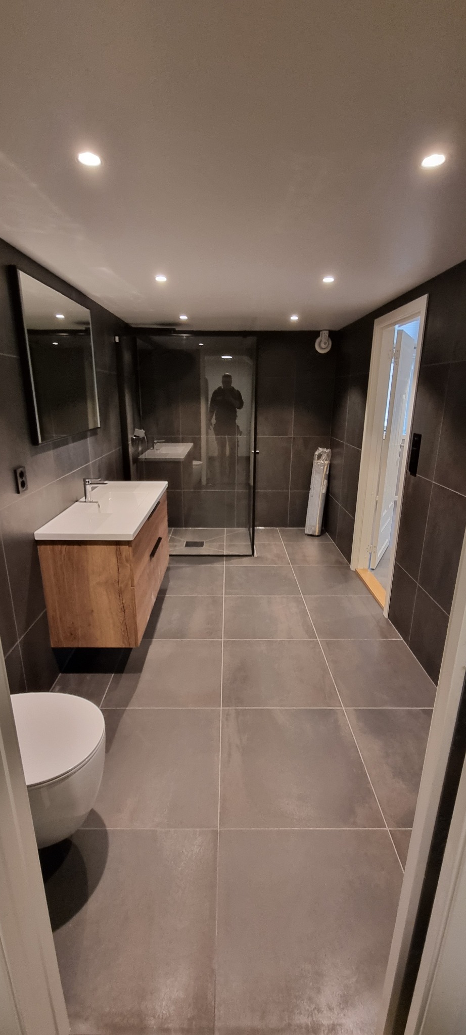 Nowoczesna łazienka z szarymi płytkami, drewnianą szafką pod umywalką, kabiną prysznicową typu walk-in i lustrem na ścianie. W lustrze kabiny widoczne odbicie osoby fotografującej.