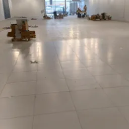 Jasna, duża przestrzeń z błyszczącą, świeżo umytą podłogą z płytek. W tle kartony na paletach i ekipa sprzątająca. Widoczne refleksy światła na podłodze.