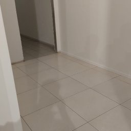 ANS Clean Craft Sprzątanie Profesjonalne i Ozonowanie - Wnętrze po remoncie: jasne płytki podłogowe i świeżo pomalowane ściany w odcieniach bieli. Widoczne przejście do kolejnego pomieszczenia.