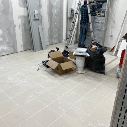 ANS Clean Craft Sprzątanie Profesjonalne i Ozonowanie - Salon w trakcie prac wykończeniowych po remoncie: podłoga z resztkami zaprawy, ściany przygotowane do malowania, instalacje elektryczne w trakcie montażu, drabina.