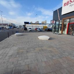 ANS Clean Craft Sprzątanie Profesjonalne i Ozonowanie - Czysta kostka brukowa przed budynkiem galerii handlowej w słoneczny dzień. Widoczne ławki i zaparkowane samochody w tle. Perspektywa z poziomu chodnika.