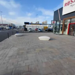 Czysta kostka brukowa przed budynkiem galerii handlowej w słoneczny dzień. Widoczne ławki i zaparkowane samochody w tle. Perspektywa z poziomu chodnika.