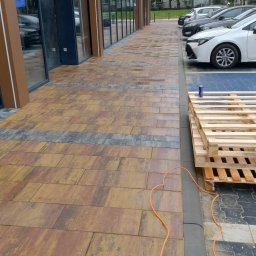 ANS Clean Craft Sprzątanie Profesjonalne i Ozonowanie - Umyta kostka brukowa przed budynkiem galerii handlowej w Skierniewicach. Widoczne palety i kabel. Kostka w odcieniach brązu i szarości.