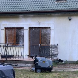 ANS Clean Craft Sprzątanie Profesjonalne i Ozonowanie - Elewacja domu w trakcie czyszczenia myjką ciśnieniową Karcher. Piana spływa po ścianach i oknach. Widoczny fragment balkonu z ozdobną balustradą.