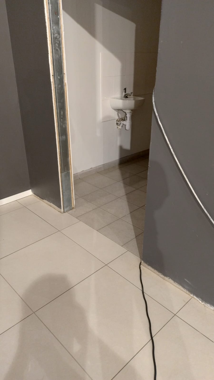 Łazienka w trakcie remontu: widoczna umywalka, płytki podłogowe i ściany w szarym kolorze. Otwarta rama drzwiowa ukazuje fragment kolejnego pomieszczenia.