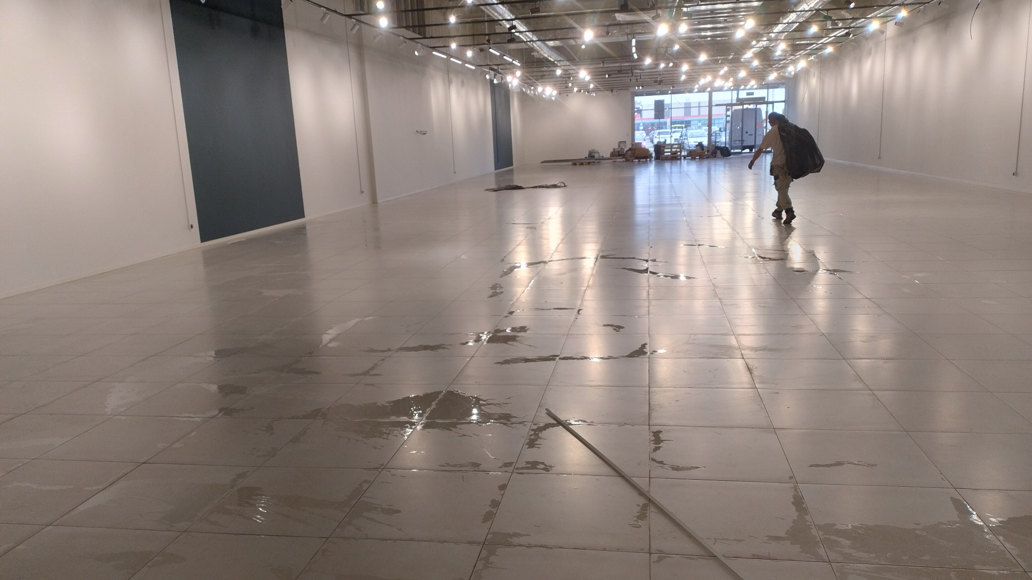 Mokry, wykafelkowany parkiet w pustym lokalu po zalaniu. W oddali osoba z workiem. Widoczne ślady wody i odbicia światła. Oświetlenie sufitowe.