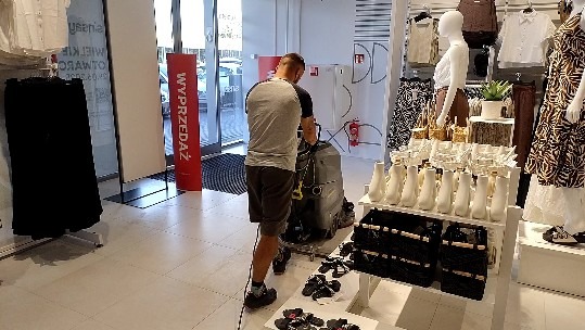 Pracownik w sklepie czyści podłogę za pomocą maszyny czyszczącej. Widoczny asortyment sklepowy i szyld 'Wyprzedaż'.