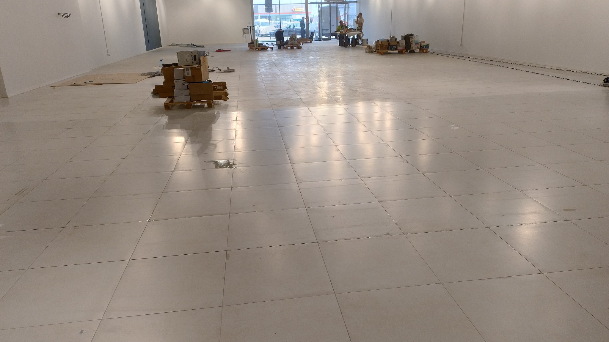 Jasna, duża przestrzeń z błyszczącą, świeżo umytą podłogą z płytek. W tle kartony na paletach i ekipa sprzątająca. Widoczne refleksy światła na podłodze.