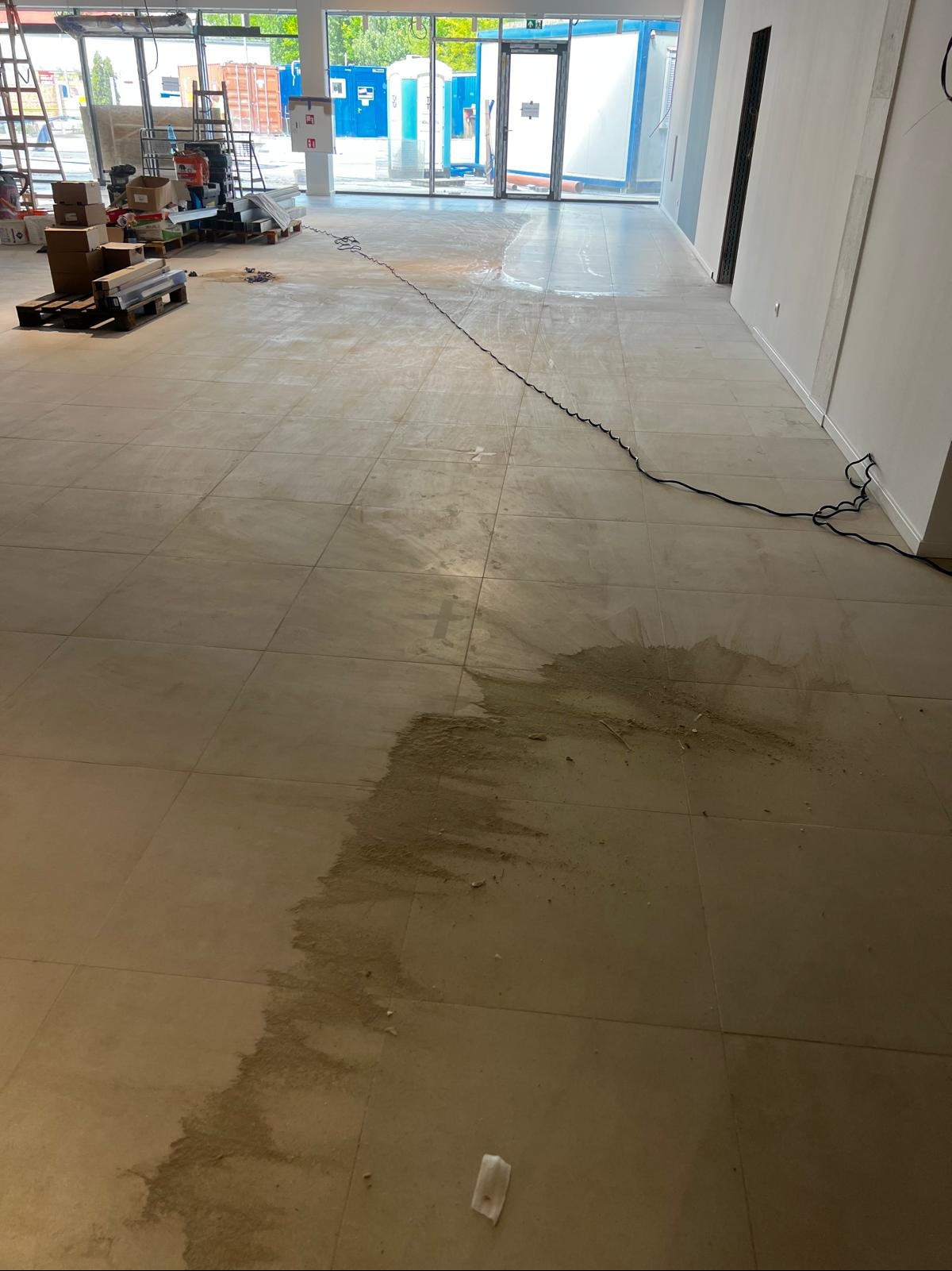 Podłoga w nowym lokalu po budowie, widoczne zabrudzenia po pracach wykończeniowych. Duże okna z widokiem na zewnątrz. Część podłogi jest mokra.