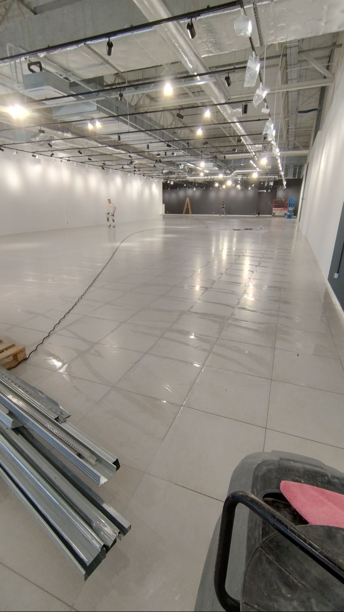 Błyszcząca, jasna podłoga w dużym, pustym pomieszczeniu po umyciu. Widoczne narzędzia i osoba w oddali. Perspektywa z dołu.