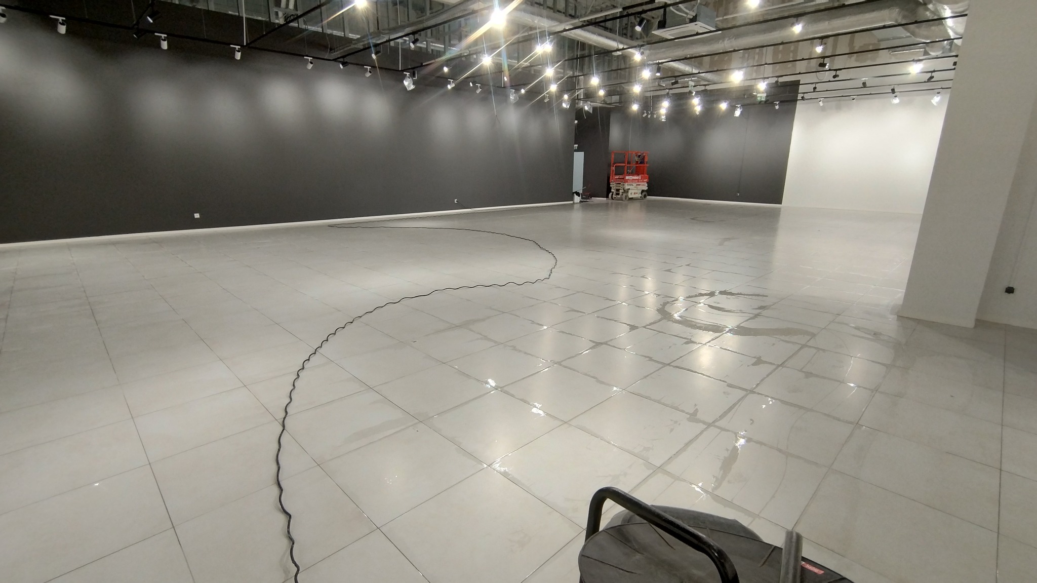 Mycie podłogi w przestronnym, nowoczesnym lokalu z czarną ścianą i białymi płytkami. Widoczne ślady wody po czyszczeniu i sprzęt do mycia podłóg w kadrze.