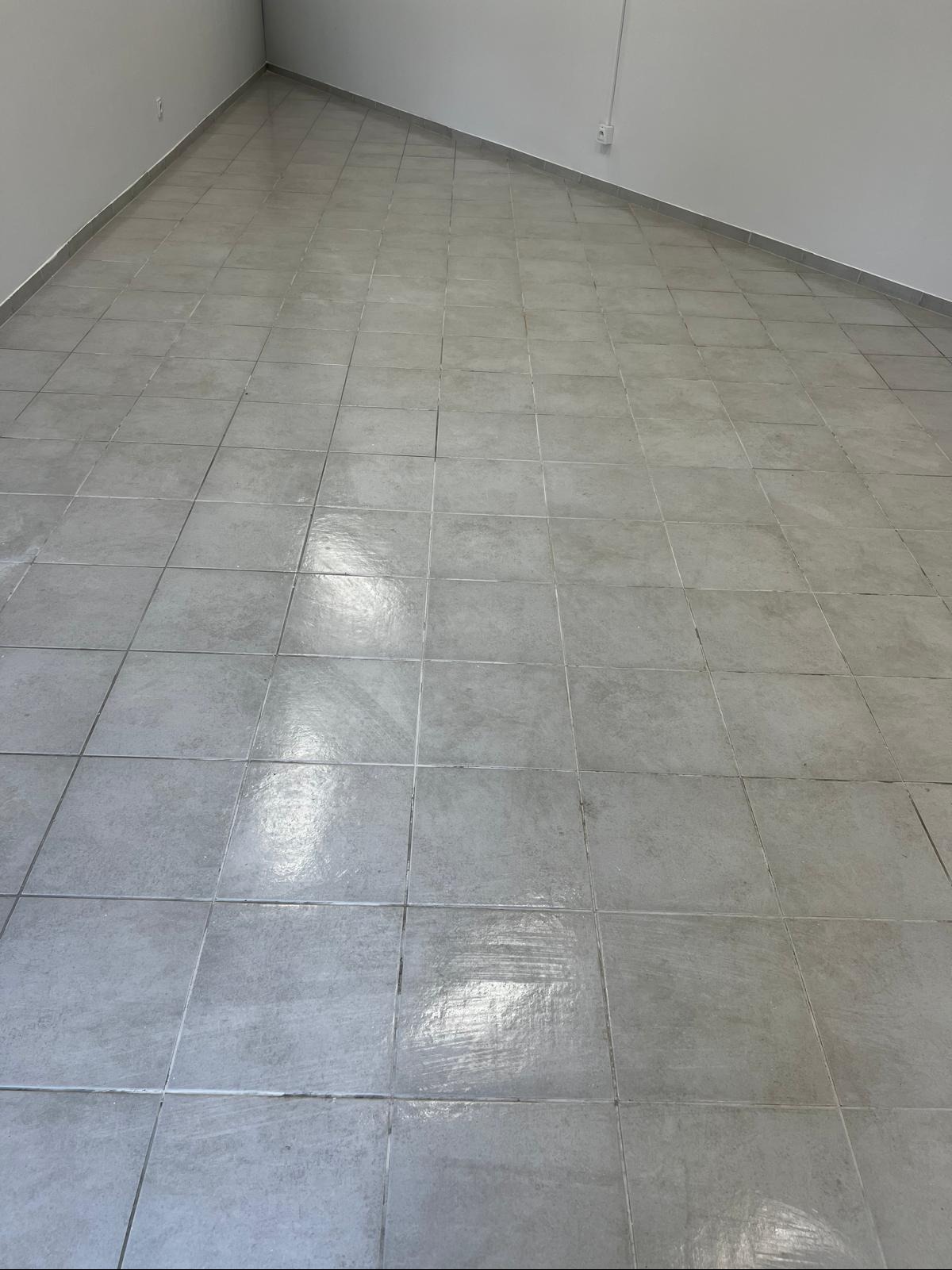 Wnętrze z szarymi płytkami podłogowymi ułożonymi w kwadraty, widoczne ślady po umyciu, ściany jasne, minimalistyczne wykończenie.