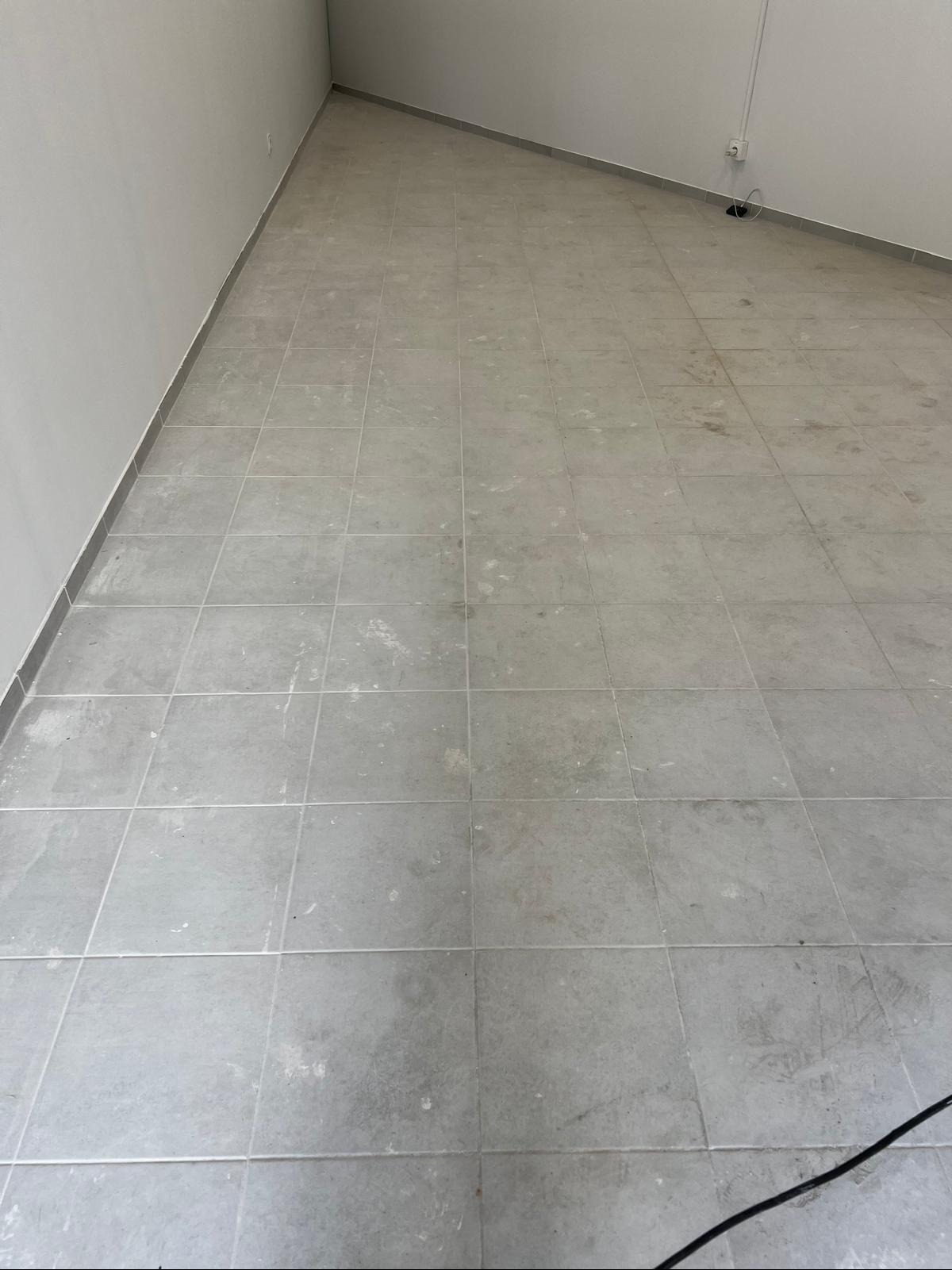 Szare płytki podłogowe w pustym pomieszczeniu po remoncie, widoczne zabrudzenia i resztki po budowie. Ujęcie z góry, kabel w rogu.