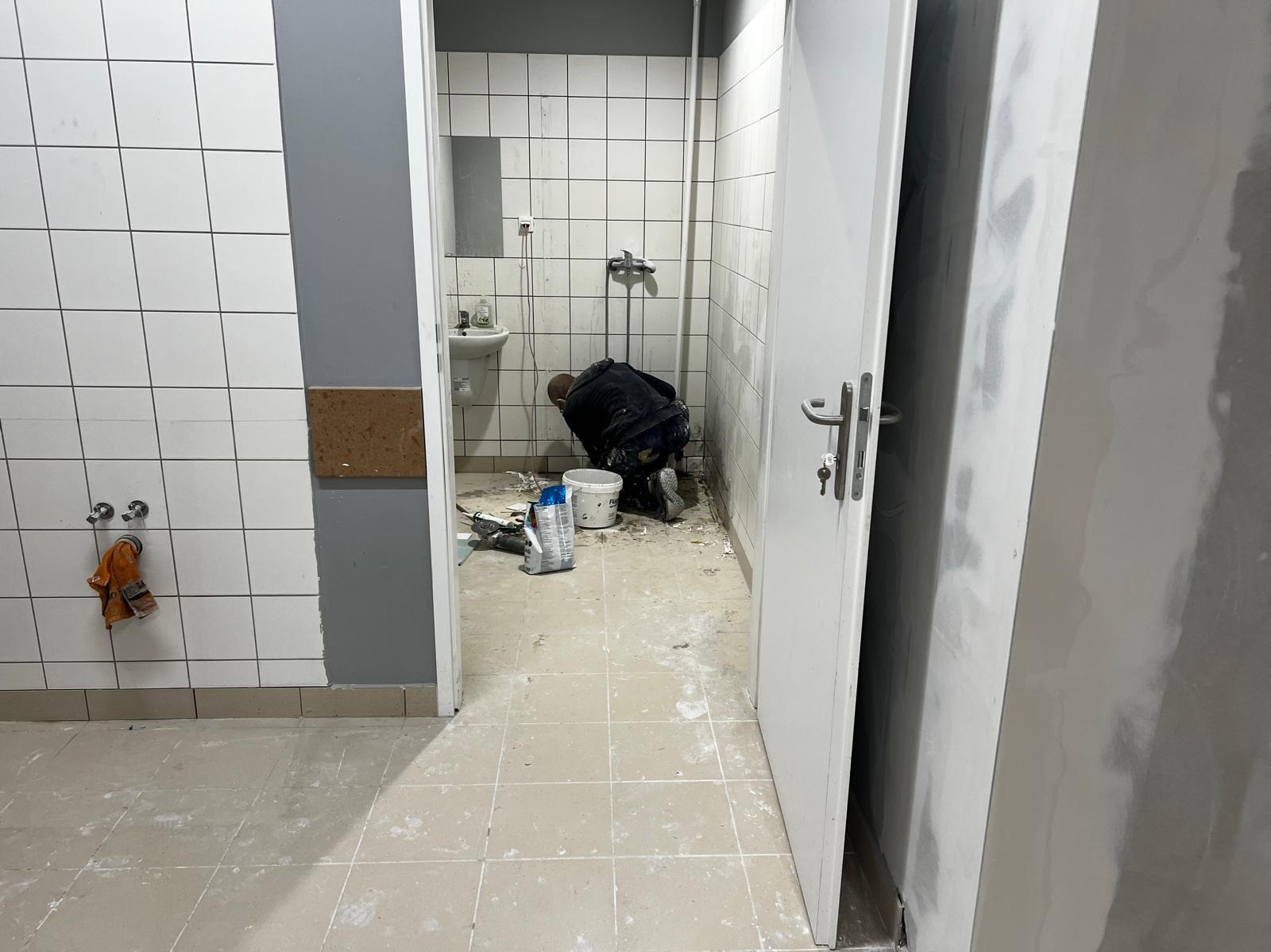 Pracownik w trakcie sprzątania łazienki po remoncie w lokalu handlowym. Widoczne narzędzia i pozostałości po pracach budowlanych na podłodze z płytek.