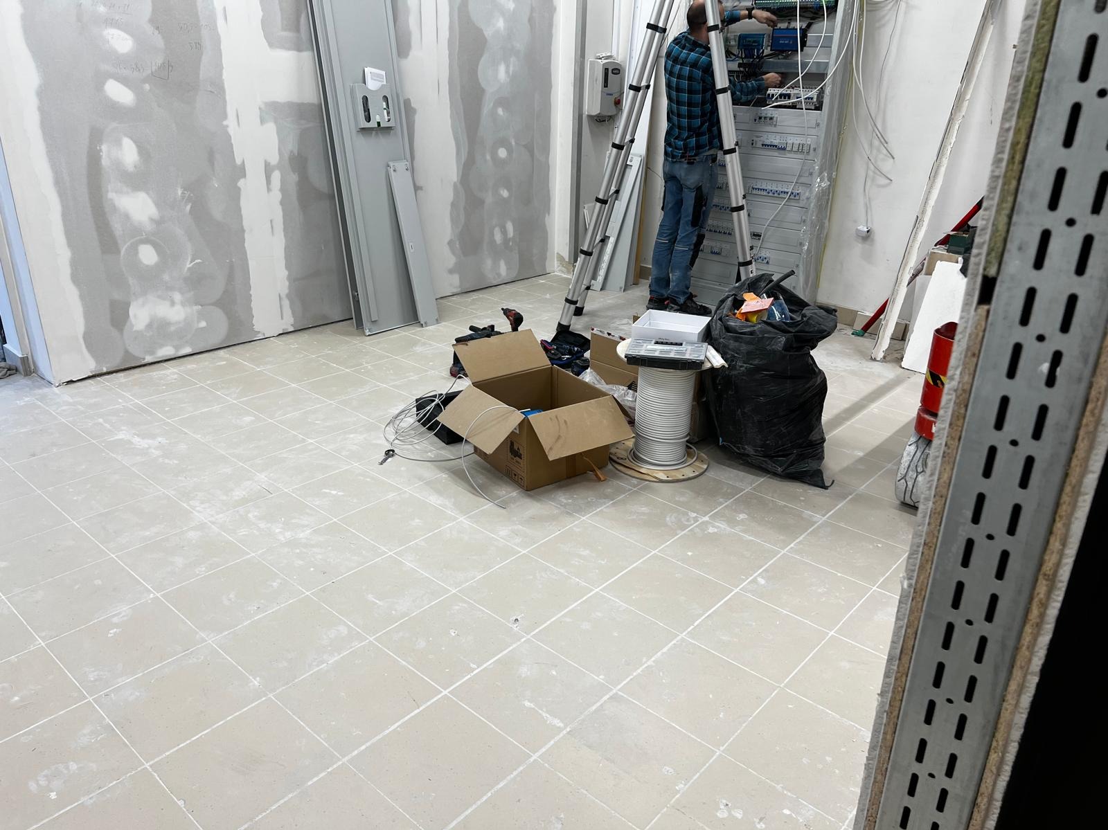 Salon w trakcie prac wykończeniowych po remoncie: podłoga z resztkami zaprawy, ściany przygotowane do malowania, instalacje elektryczne w trakcie montażu, drabina.