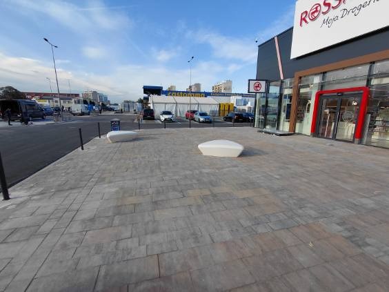 Czysta kostka brukowa przed budynkiem galerii handlowej w słoneczny dzień. Widoczne ławki i zaparkowane samochody w tle. Perspektywa z poziomu chodnika.