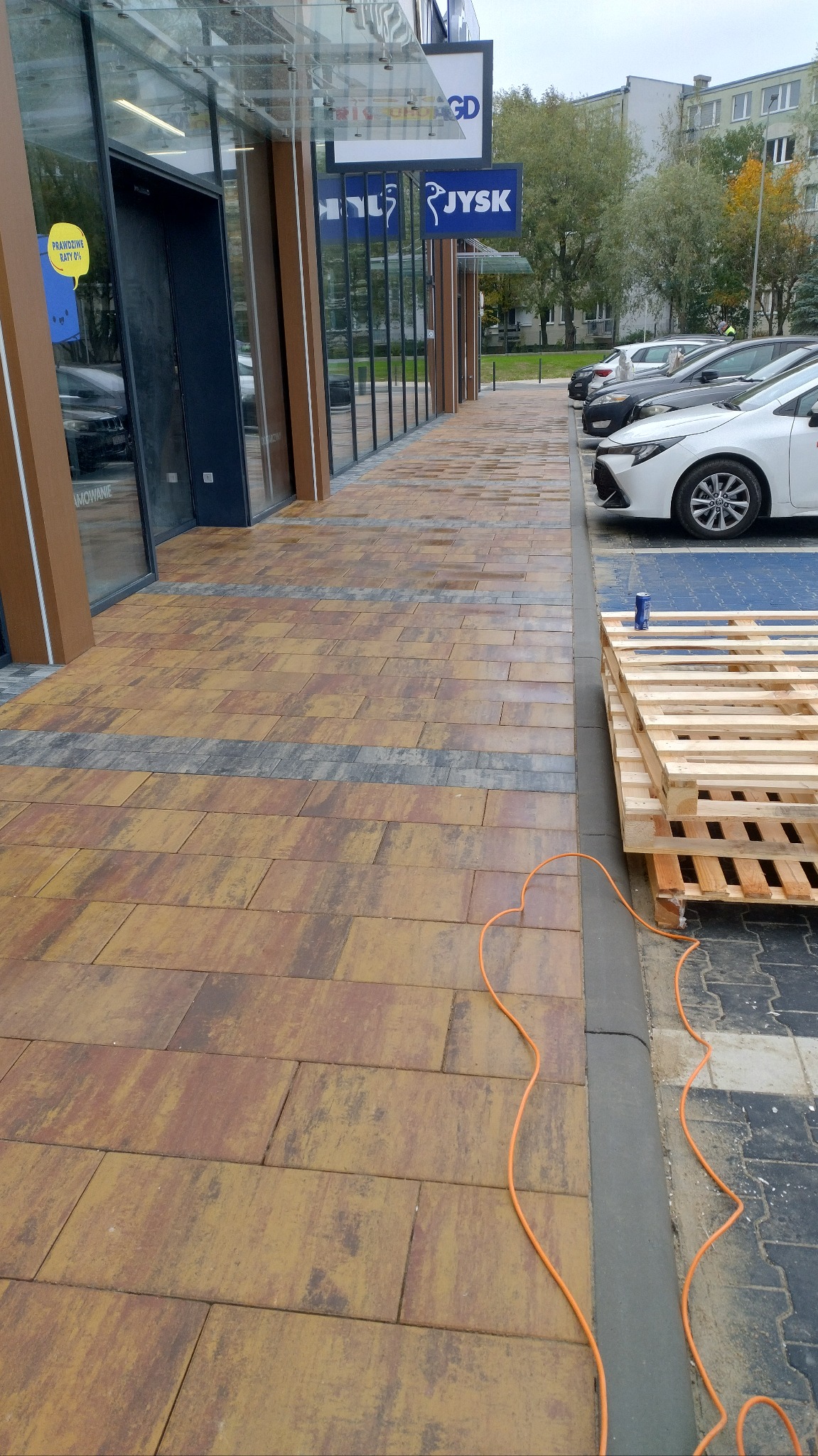 Umyta kostka brukowa przed budynkiem galerii handlowej w Skierniewicach. Widoczne palety i kabel. Kostka w odcieniach brązu i szarości.
