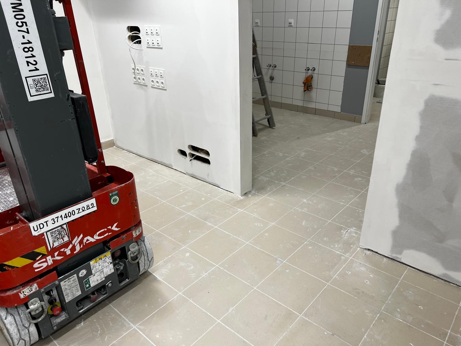 Salon po remoncie w galerii handlowej: podłoga w płytkach z resztkami farby, ściany z gniazdkami, podnośnik Skyjack. Widok na pomieszczenie z drabina i umywalkami.