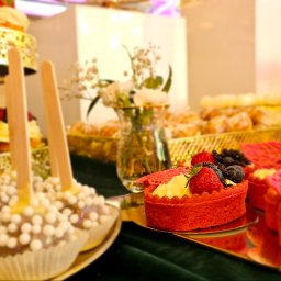 Rezydencja - Bogato zastawiony stół z deserami: czerwone tartaletki z owocami, cake pops z białymi kuleczkami, babeczki i inne słodkości na złotych paterach, w tle wazon z białymi kwiatami.