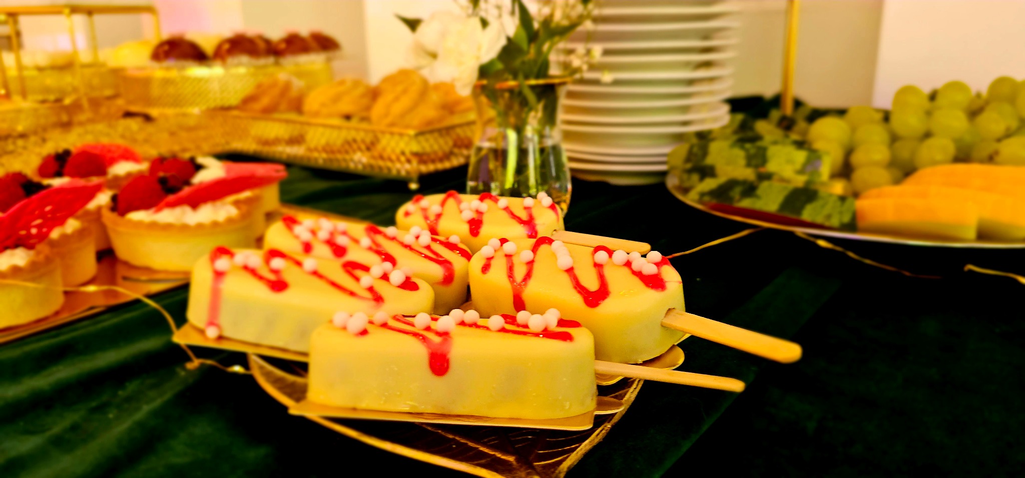 Elegancki stół cateringowy z deserami: mini tartaletki z truskawkami, ciasteczka, owoce i lody na patyku z białą polewą, czerwonym sosem i białymi kuleczkami, ustawione na zielonym obrusie.