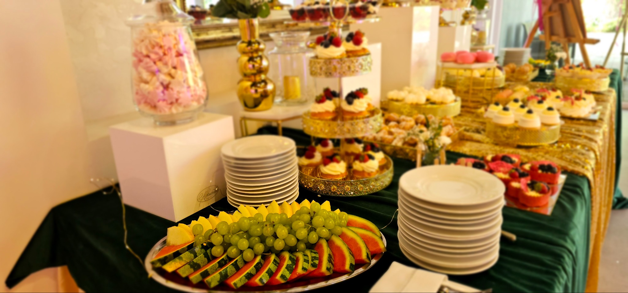 Stół z przekąskami: owoce (arbuz, winogrona, mango) na srebrnej tacy, stosy białych talerzy, babeczki z owocami na złotych paterach, słoik z piankami, złota waza, zielony obrus, złoty bieżnik.