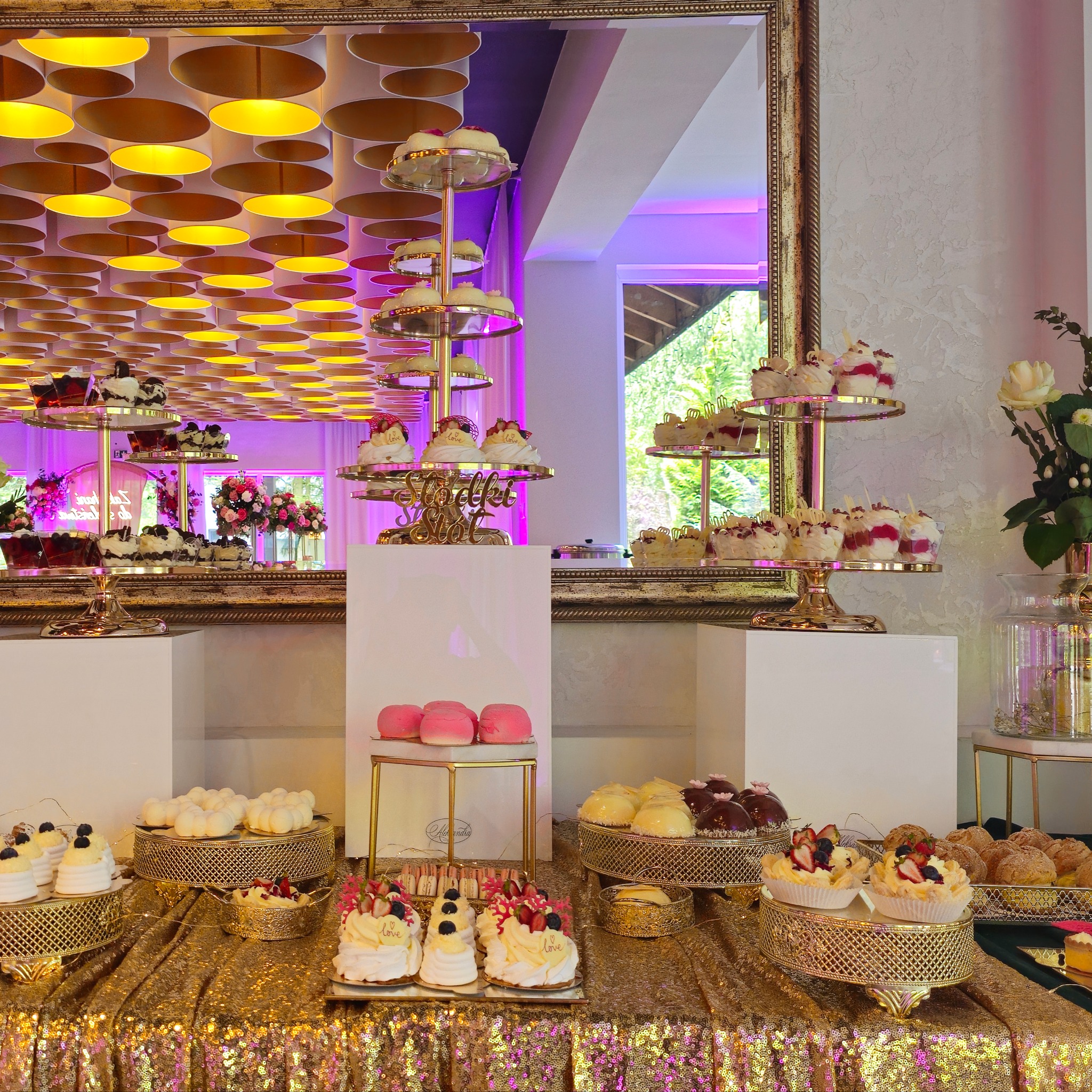 Bogato zastawiony stół z różnorodnymi deserami na złotym, błyszczącym obrusie; w tle lustro odbijające dekoracje kwiatowe i nowoczesne oświetlenie sufitowe.