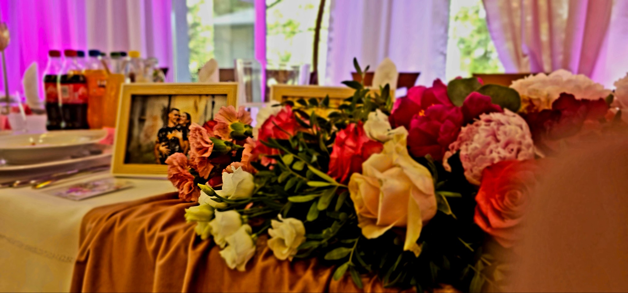 Dekoracja stołu z bukietem róż, goździków i zielonych liści, ramki ze zdjęciami pary, napoje w tle i nakrycia na stole podczas uroczystości.