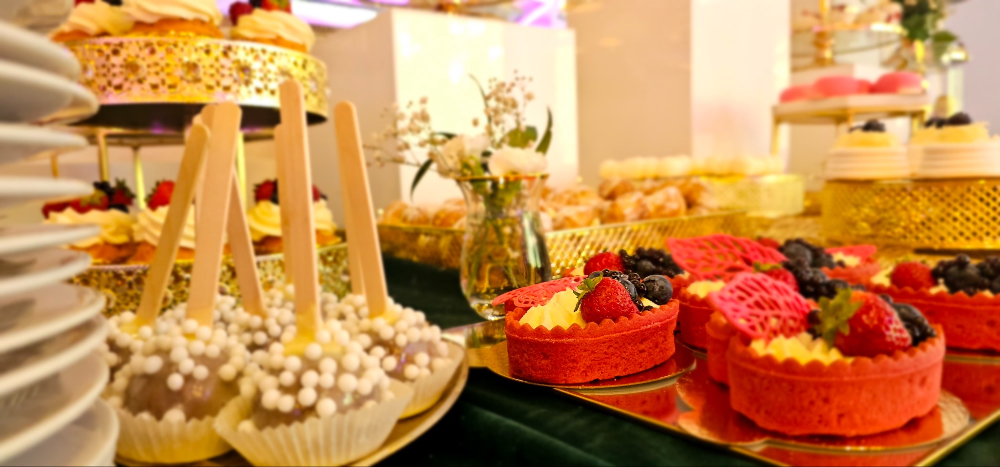 Bogato zastawiony stół z deserami: czerwone tartaletki z owocami, cake pops z białymi kuleczkami, babeczki i inne słodkości na złotych paterach, w tle wazon z białymi kwiatami.