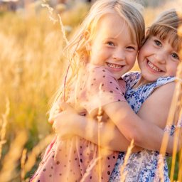 Pan Aparacik Dawid Kubica - Dwie uśmiechnięte dziewczynki obejmują się na tle pola zboża w ciepłym, zachodzącym słońcu, jedna w różowej sukience w kropki, druga w niebieskiej w kwiaty.